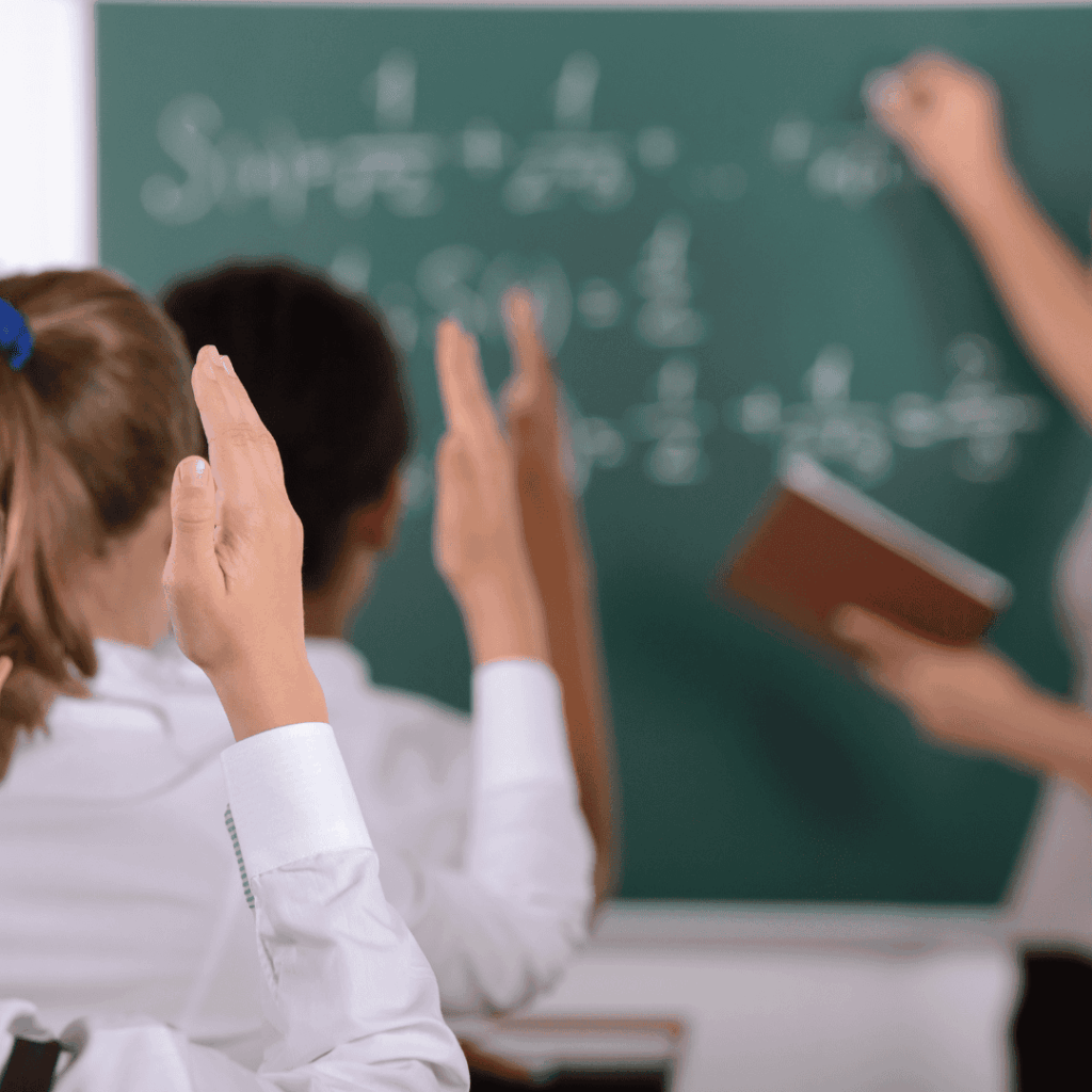 Uczniowie w klasie podnoszą ręce do odpowiedzi, nauczyciel pisze wzory matematyczne na tablicy.