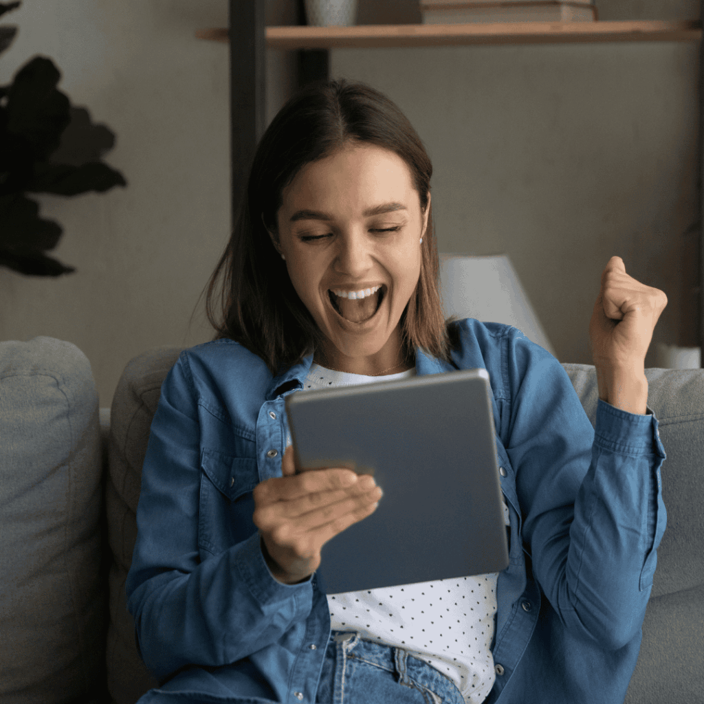 Uśmiechnięta kobieta siedząca na kanapie, cieszy się patrząc na tablet i unosi zaciśniętą pięść w geście radości.